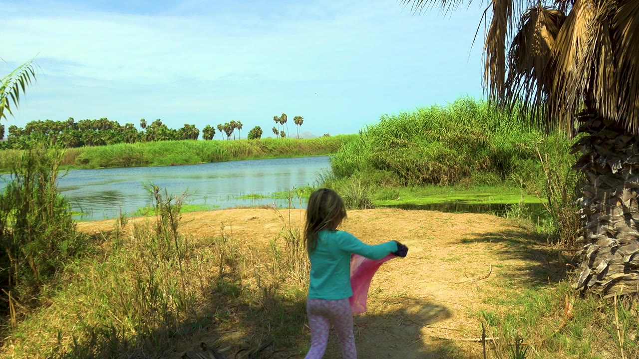 A young girl in a turquoise top and pale pink pants walks away from camera in San Jose del Cabo, holding a pink cloth near water, reeds, palms, and dry ground.