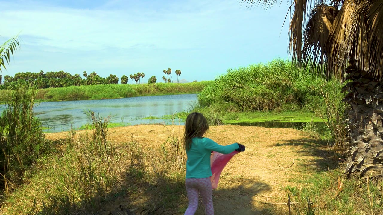 The girl stands farther into the clearing in San Jose del Cabo, facing away while holding the pink cloth, with water to the left and dense green grasses ahead.