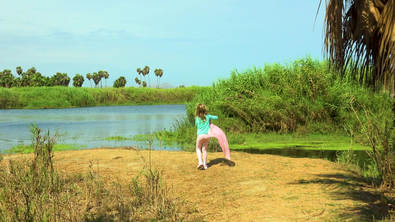 In San Jose Del Cabo, Mexico, a young blonde girl stands on a sandy bank with a pink cloth beside blue water, green reeds, and distant palms.