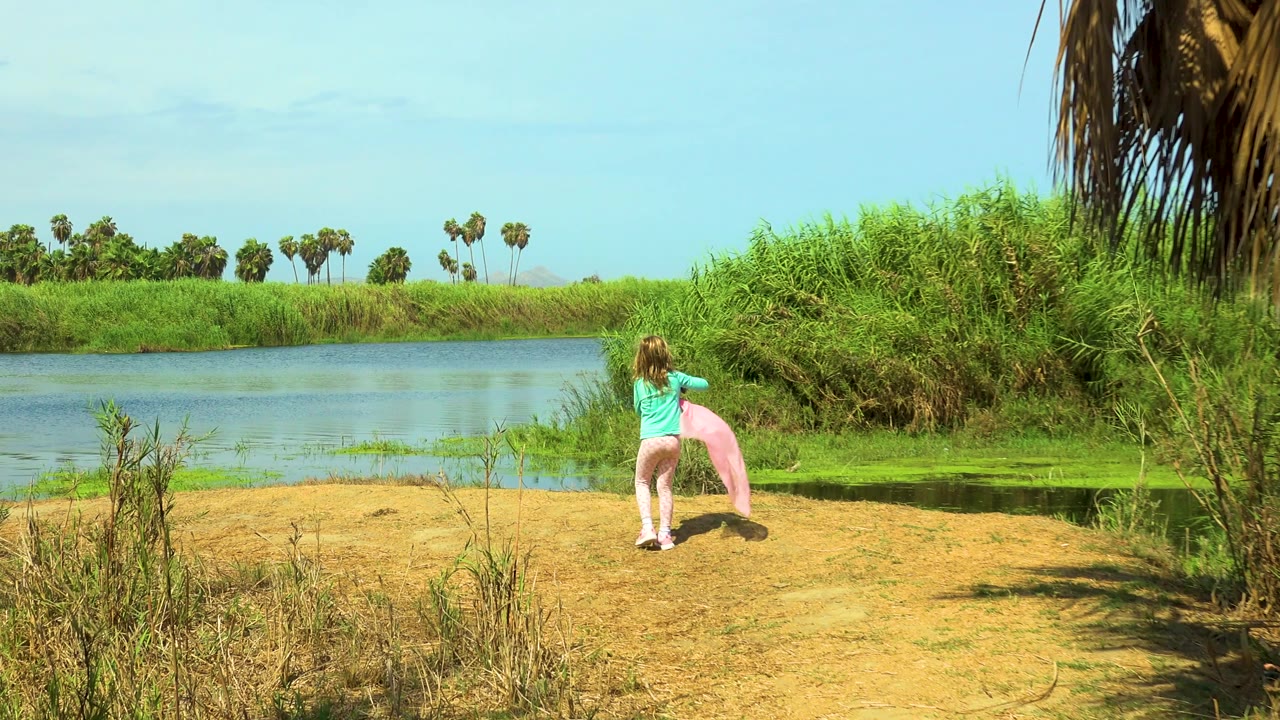 Young girl facing water with a pink cloth on a sandy shore.