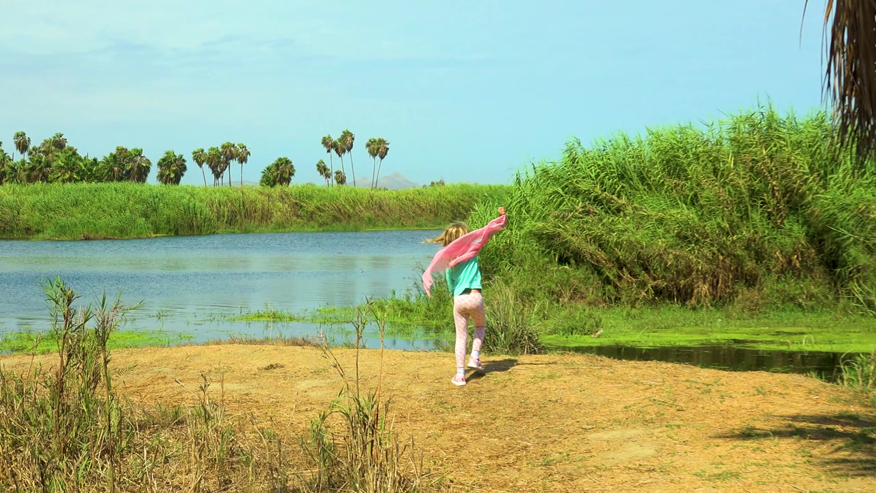 In San Jose Del Cabo, Mexico, the young girl lifts the pink cloth to one side near the water, with dense green reeds and palms under bright daylight.