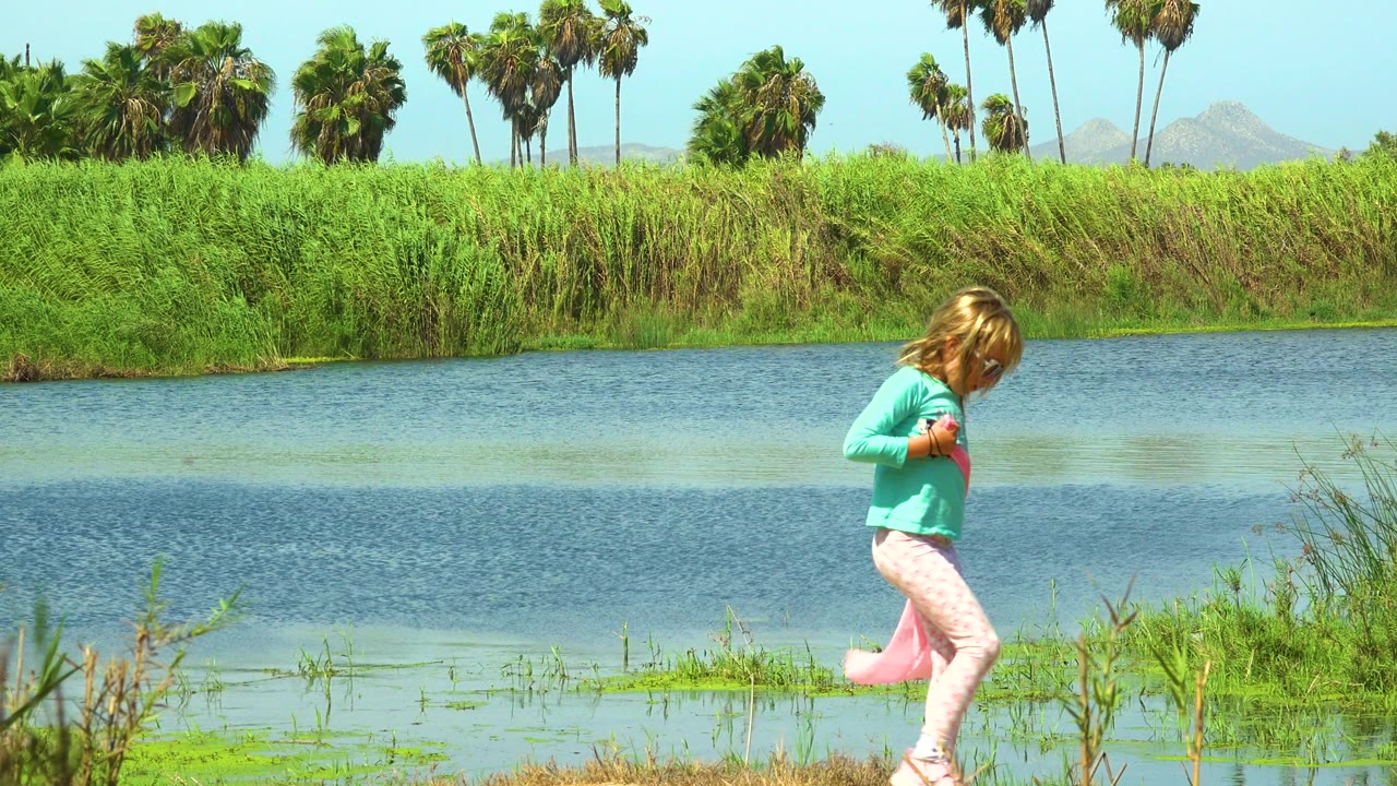 Young girl in a turquoise top walks along the San Jose Del Cabo riverbank with a pink cloth trailing near her legs and palm trees across the water.