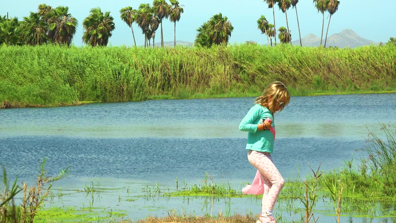Girl looks down while walking near reeds and river water.