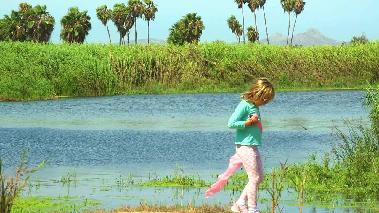 The child continues across the San Jose Del Cabo riverbank with the pink cloth stretched behind her, blue water in the midground, and palms above the reed line.