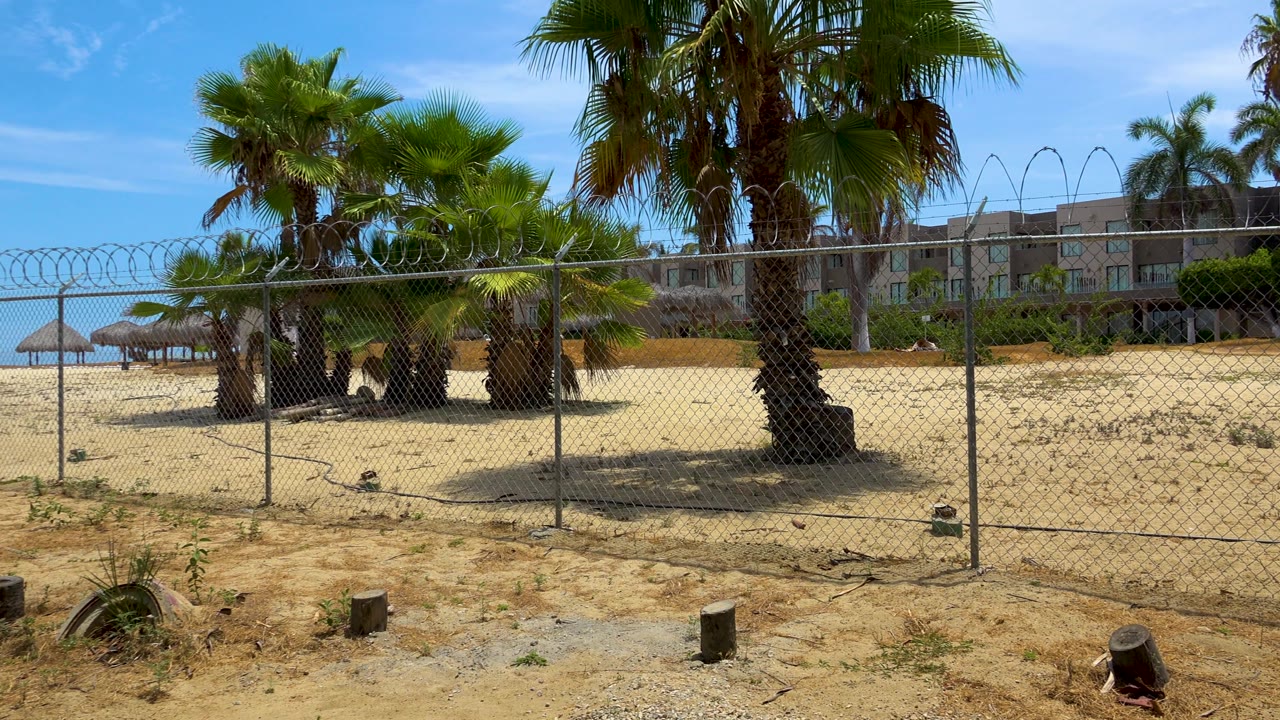 Third frame of the San Jose Del Cabo beach perimeter keeps the fence central, with clustered palms behind it, pale sand, low hotel buildings, clear blue sky, and no visible people.