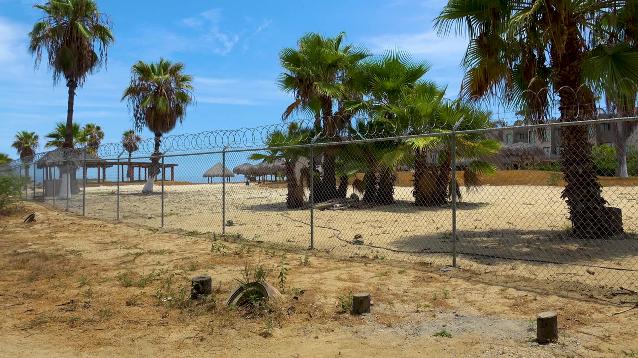 Bright midday frame in San Jose del Cabo showing dry sandy foreground, a chain-link fence, palms, thatched shade structures, a low ocean line, and no visible people.