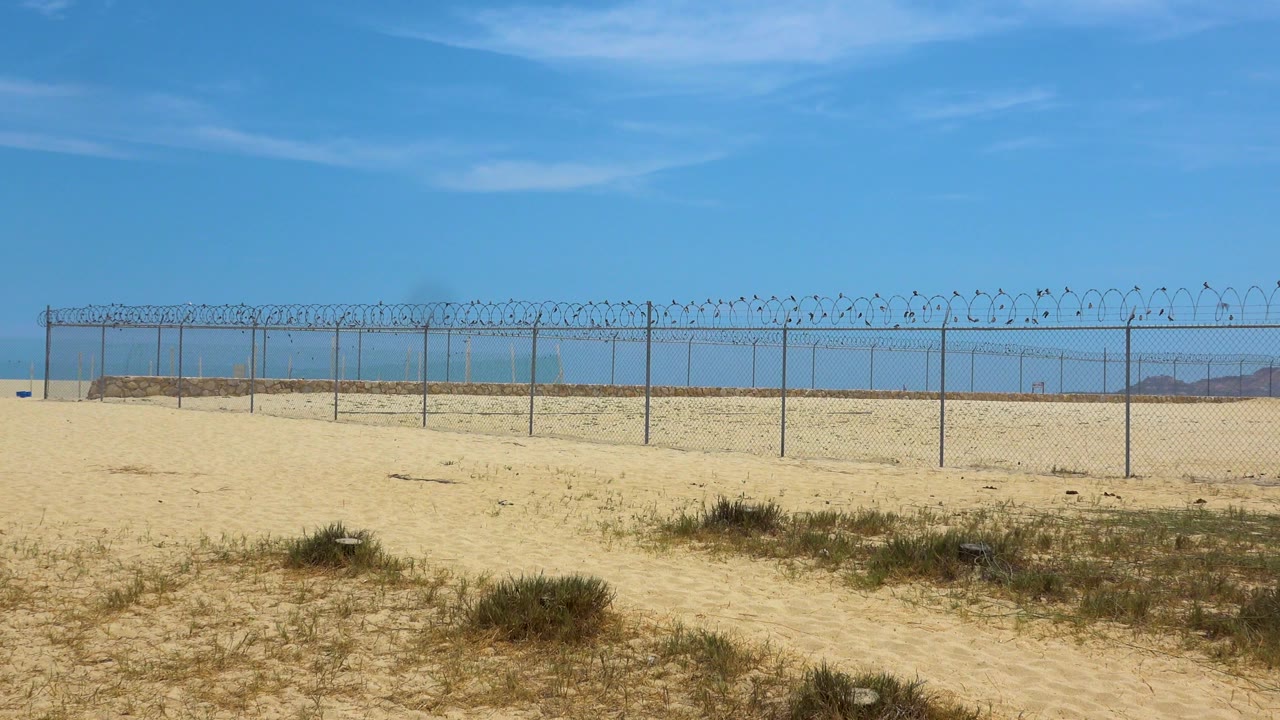 Wide ground-level view in San Jose Del Cabo, Mexico, showing sandy foreground scrub, a long chain-link fence with coiled wire, blue horizon, and no visible people.
