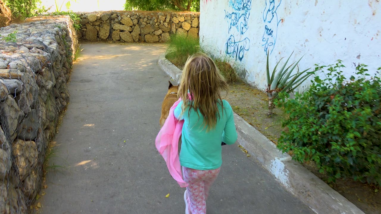 At 0:02, the child holds a pink blanket while walking behind the dog near a white wall marked with blue graffiti and low greenery in San Jose del Cabo, Mexico.