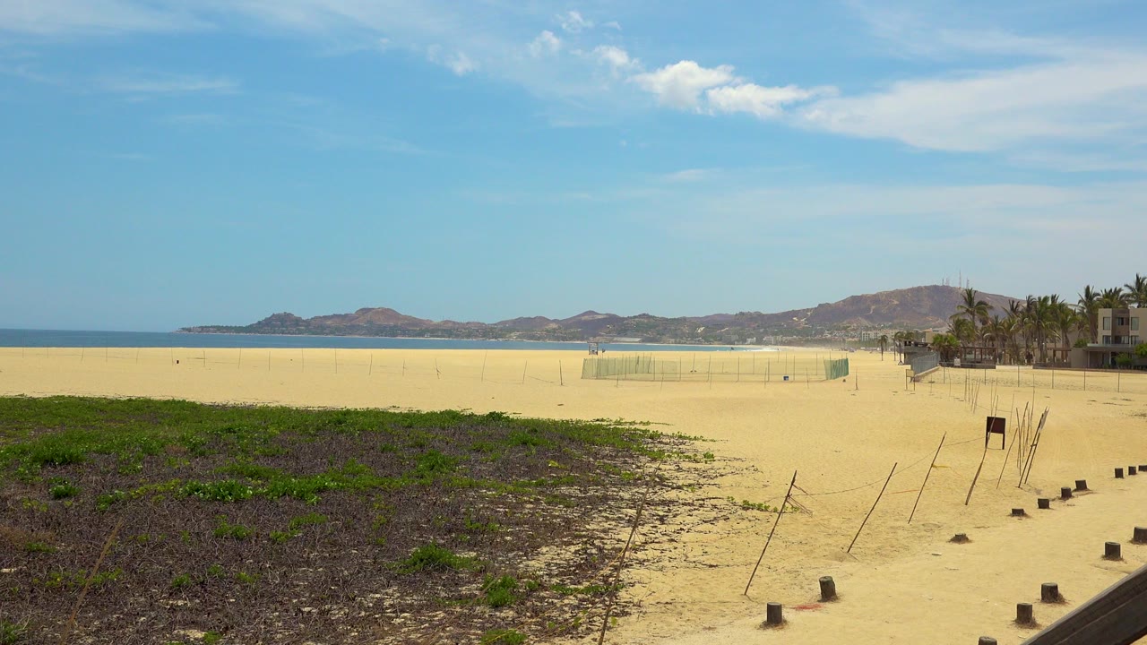 Opening frame of San Jose del Cabo beach with low foreground vegetation, pale sand, blue ocean at left, distant mountains, and no visible people.