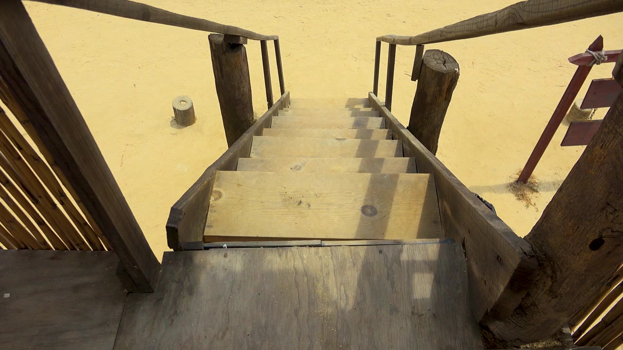 The second frame keeps the San Jose Del Cabo stairway centered, with sunlit treads, dark timber railings, pale sand below, and no visible people.