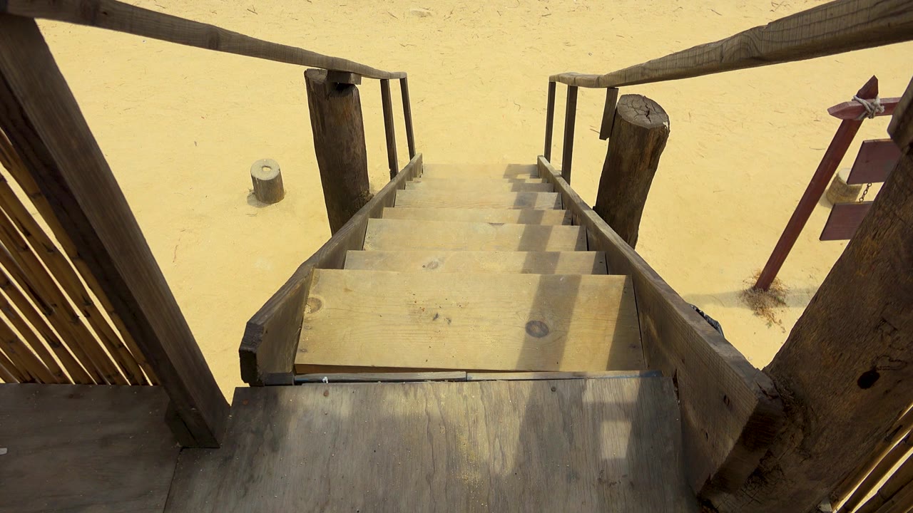 A repeated high view shows the heavy timber rails framing the beach access stairs in San Jose Del Cabo, with pale sand and a small cylindrical bollard visible, and no visible people.