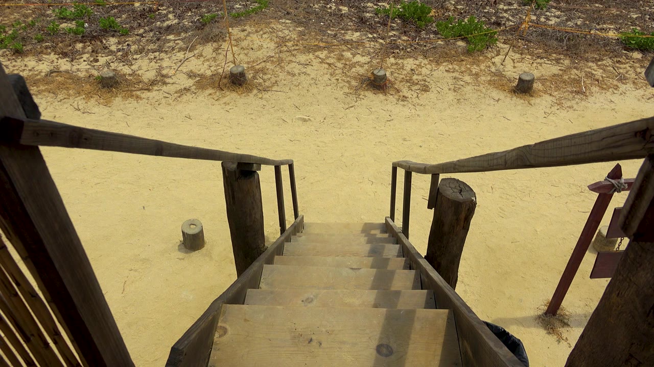 Opening view from the top of wooden stairs in San Jose Del Cabo, Mexico, with pale sand, scattered weeds, posts, and no visible people.
