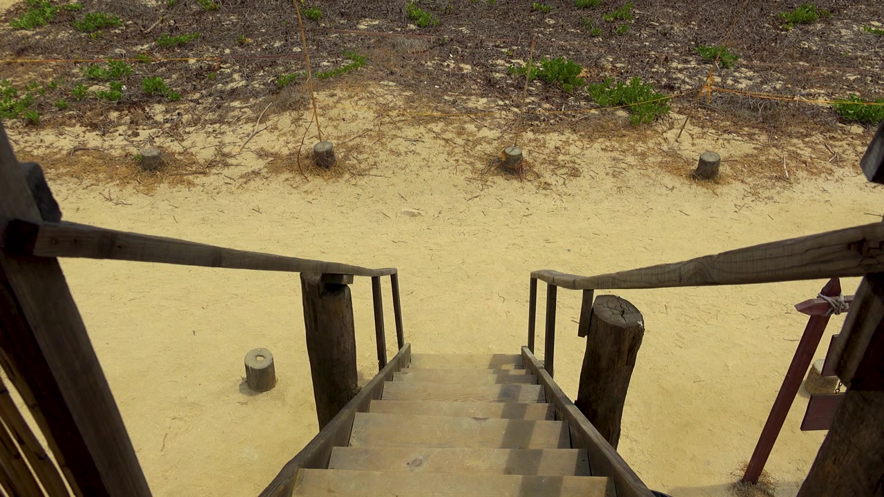 The stairway sits lower in the frame while sand, weeds, and sparse green plants fill more of the San Jose Del Cabo beach access view, with no visible people.