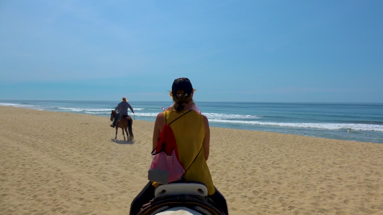 In San Jose del Cabo, Mexico, a woman in a yellow top rides a horse from behind while another rider moves ahead beside low ocean waves.