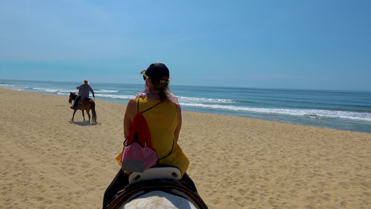 A woman seen from behind rides across the sand in San Jose del Cabo, Mexico, carrying a bright bag while the rider ahead angles closer to the water.