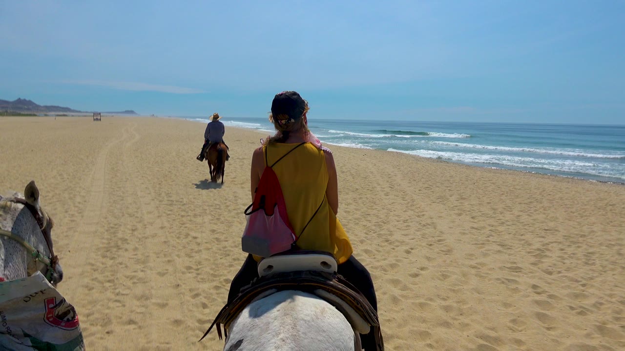 A rider in a yellow top sits on a white horse in San Jose Del Cabo, Mexico, following another rider along open sand with surf rolling on the right.