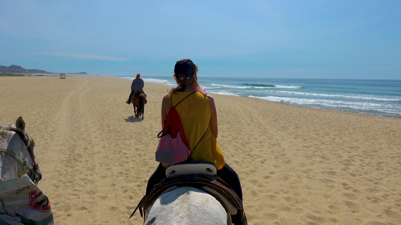 Horseback riders continue along the sandy shoreline with surf to the right.