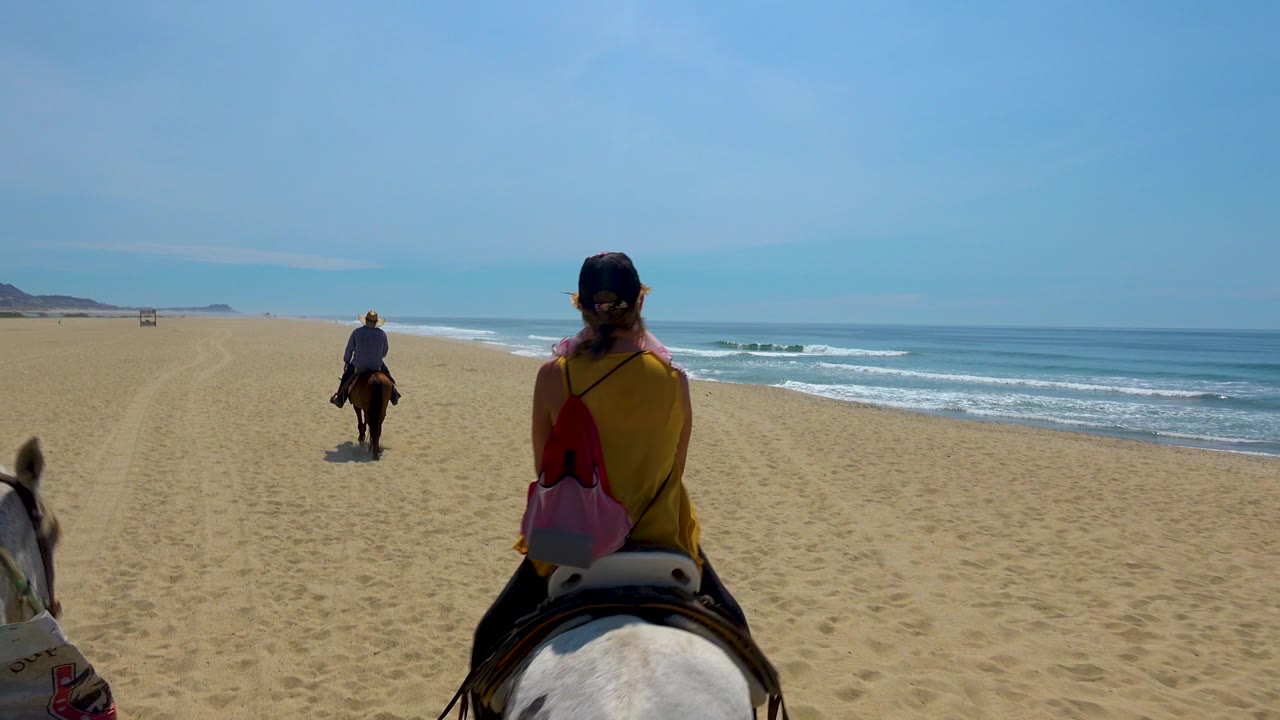 A rear view shows the nearest rider and white horse slightly right of center in San Jose Del Cabo, Mexico, with the lead rider farther up the sand and surf bands visible to the right.