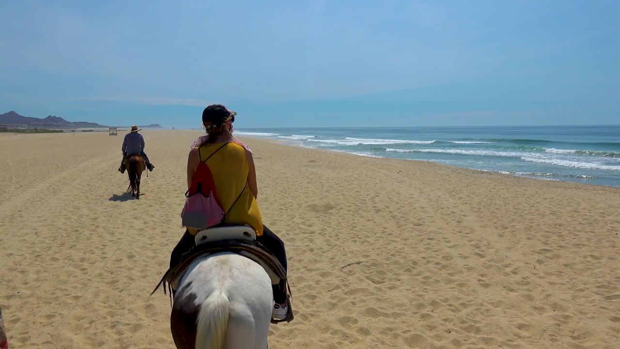 At 0:00 in San Jose del Cabo, Mexico, the camera looks past a foreground rider on a white horse toward another rider ahead on the open beach, with surf rolling along the right side.