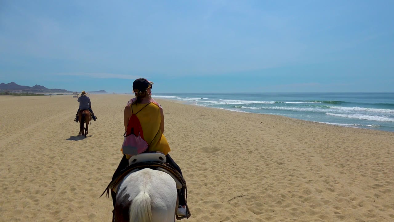 At 0:02 in San Jose del Cabo, Mexico, the foreground rider shifts slightly right in frame as the second rider stays ahead, with a broad stretch of empty sand and low mountains on the left horizon.