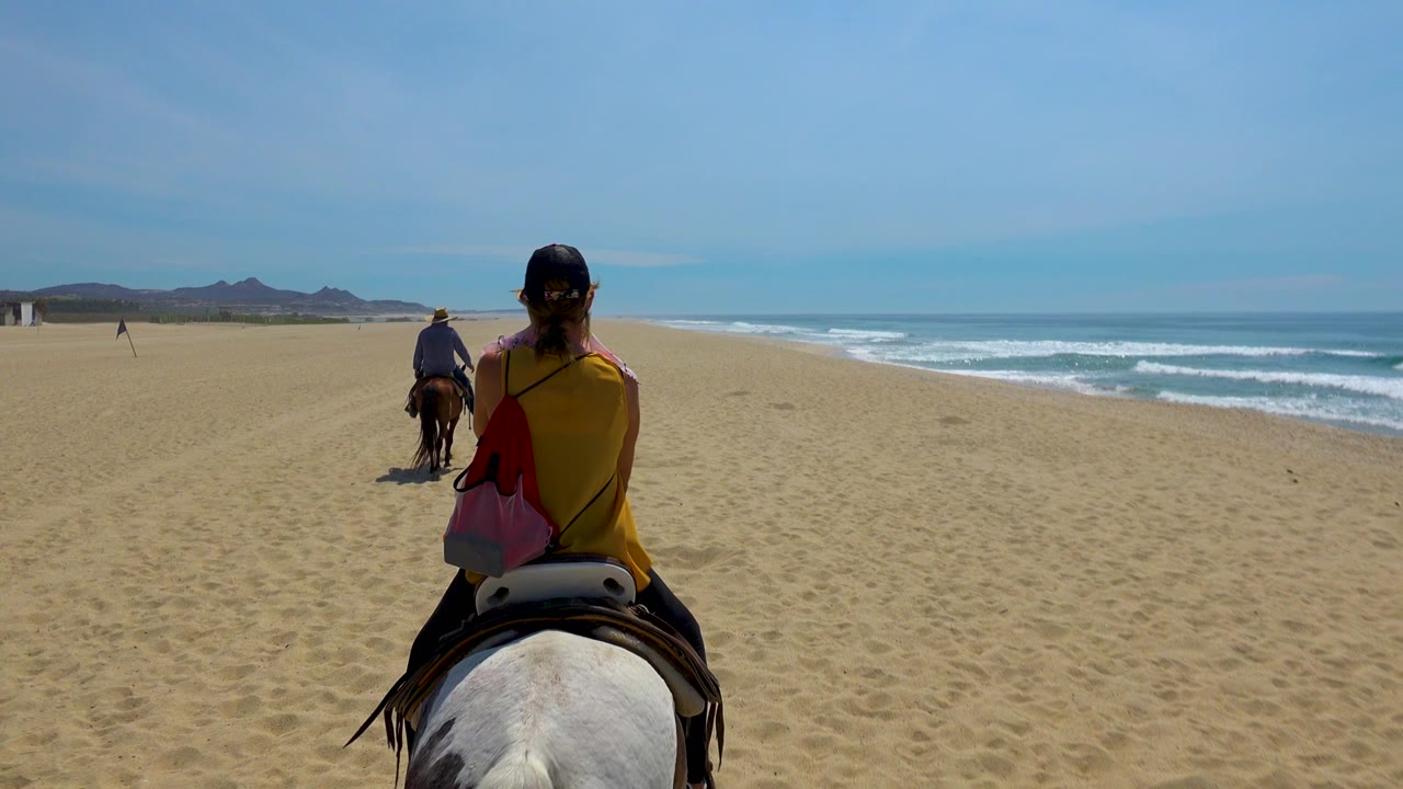 At 0:01 in San Jose Del Cabo, Mexico, the nearest rider remains centered from behind while the lead horse moves ahead across the beach and the white surf line runs along the right edge.