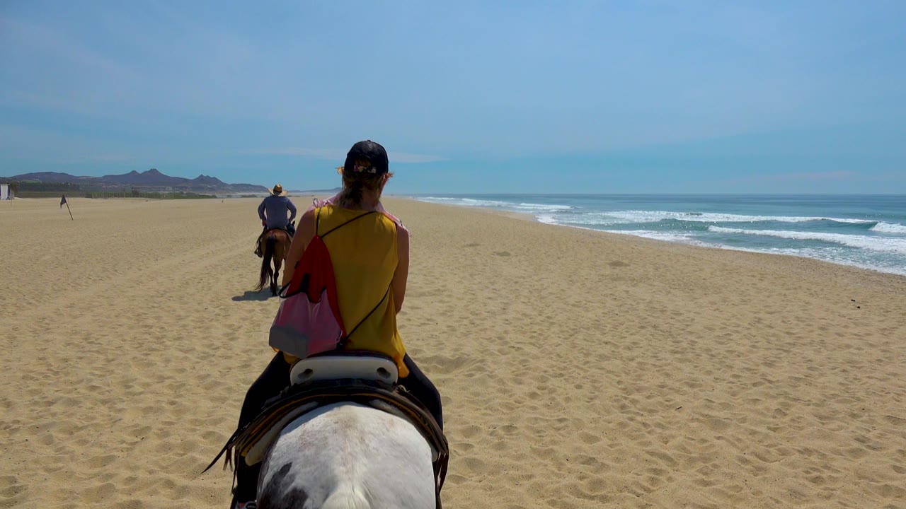 At 0:02 in San Jose Del Cabo, Mexico, the closest rider in a yellow top and cap is seen from behind, with another horse ahead, flat beach to the left, and waves breaking under a pale blue sky.