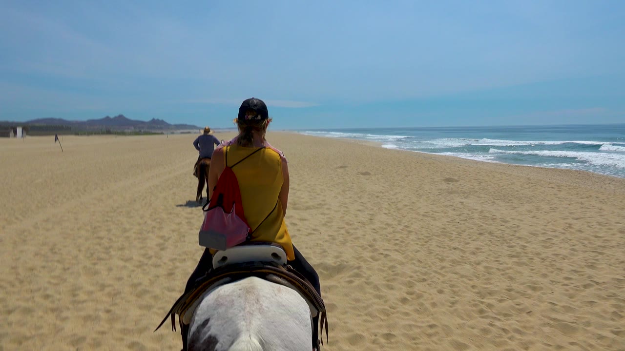 At 0:03 in San Jose Del Cabo, Mexico, the horseback line is slightly farther along the beach, with the nearest horse filling the lower frame and white waves rolling parallel to the riders.