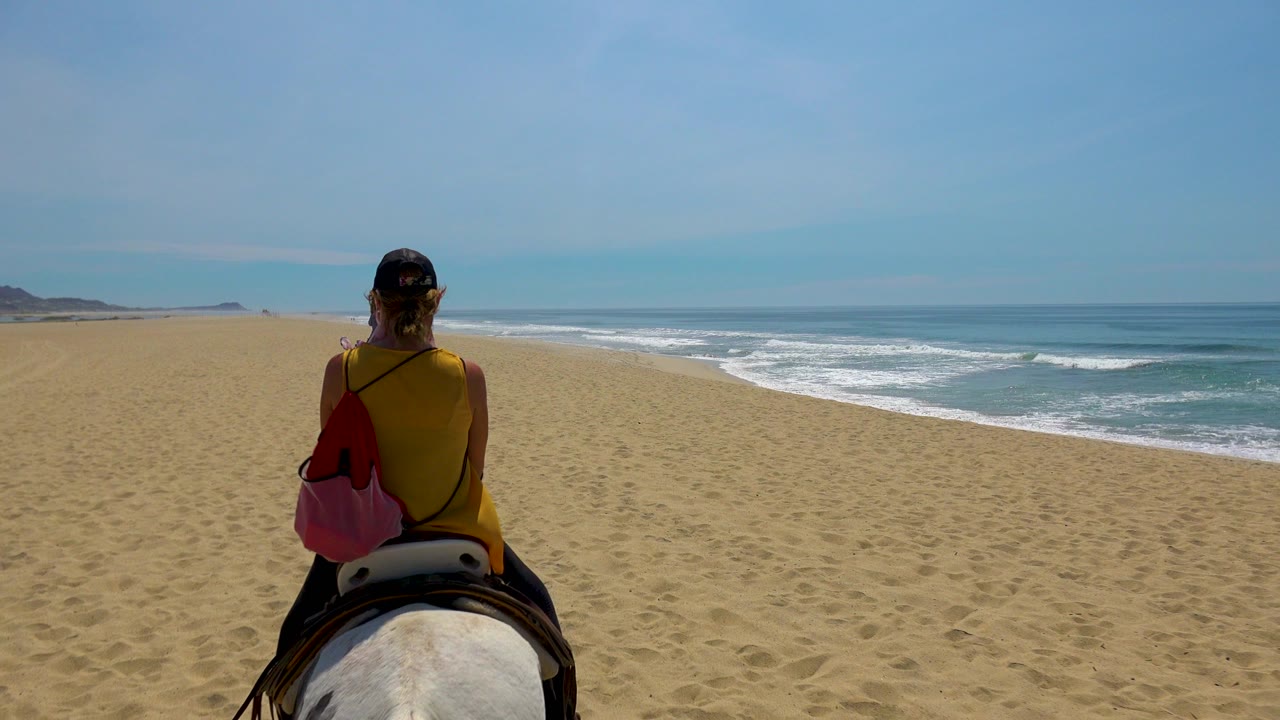 At 0:01 in San Jose Del Cabo, Mexico, the horse and rider remain centered while the shoreline stretches ahead and low waves roll in from the right.