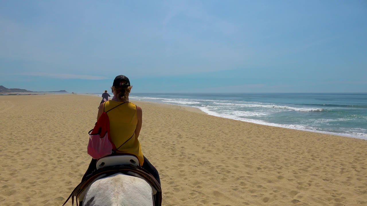At 0:02 in San Jose Del Cabo, Mexico, the rider on horseback is seen from behind, with a small distant figure on the sand and surf breaking to the right.