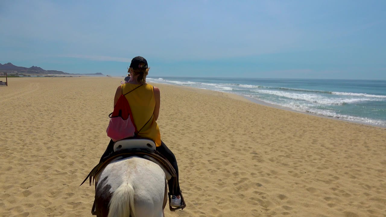 A rear view of the rider on horseback continues along the San Jose Del Cabo beach, with the ocean horizon stretching across the right side of the frame.