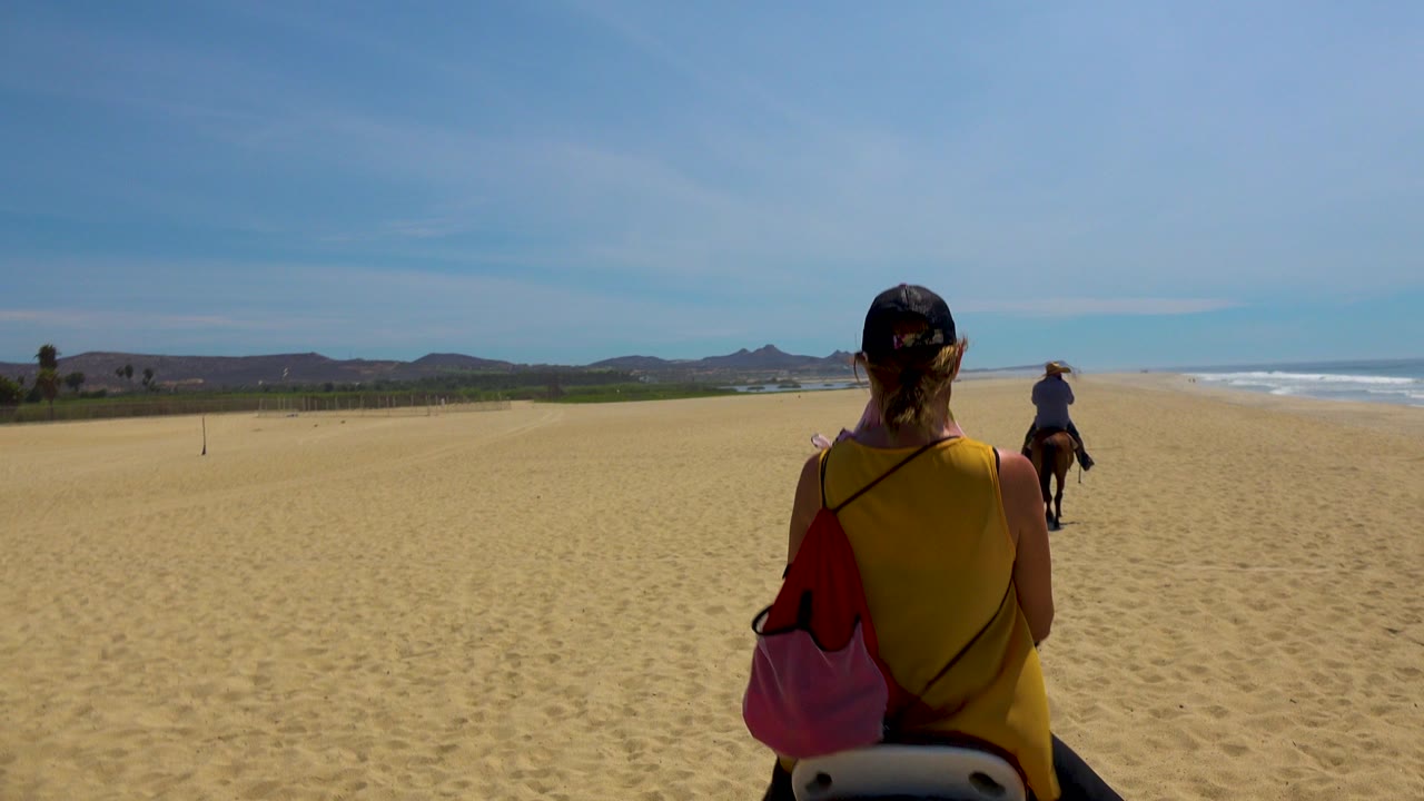 A foreground rider in a yellow top is seen from behind on horseback on a wide San Jose Del Cabo beach, with another rider ahead and the ocean on the right.