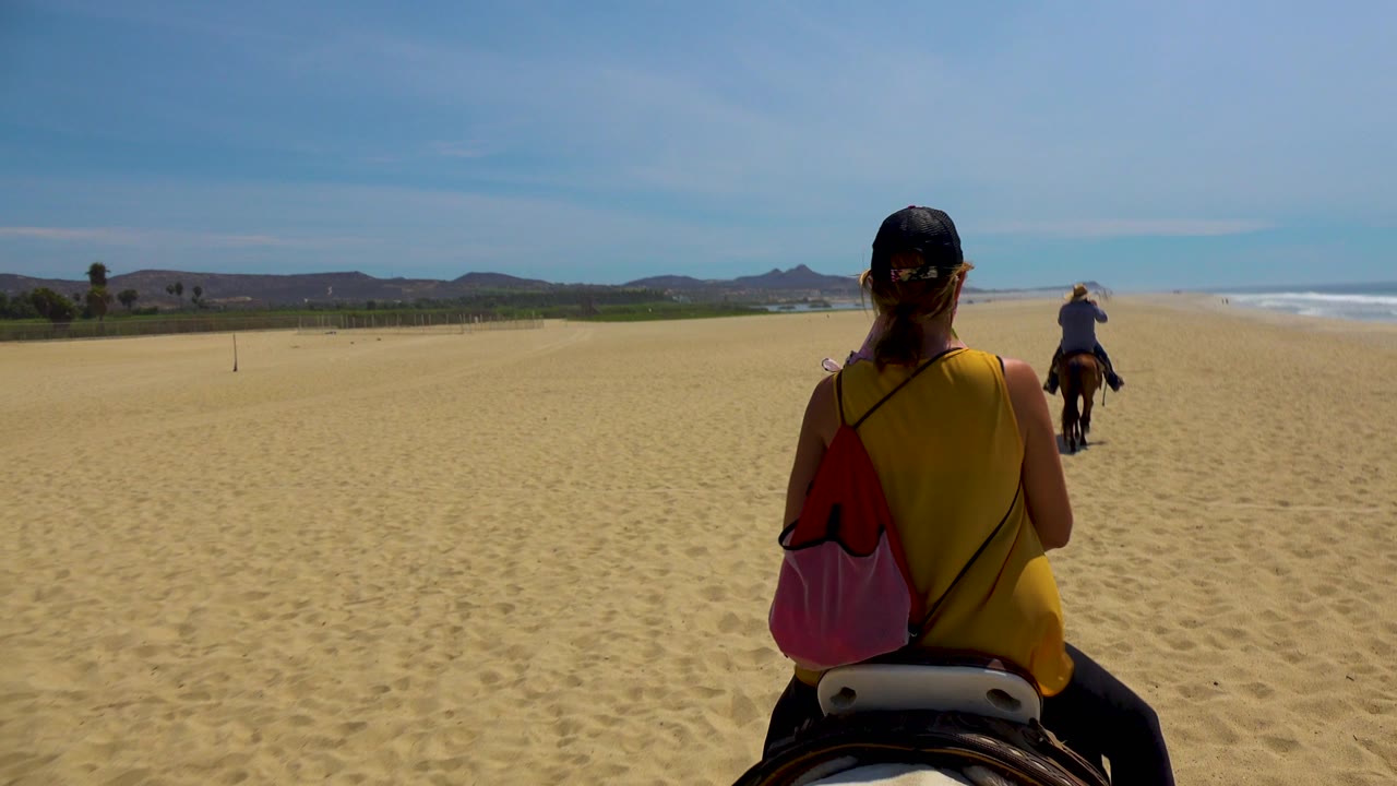 A closer rear view shows the foreground rider's cap, yellow top, and pink bag while the horse moves along the sandy San Jose Del Cabo beach near the ocean.
