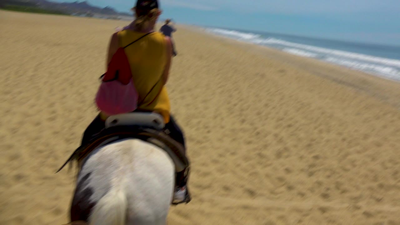 Opening frame in San Jose Del Cabo, Mexico, with a rider seen from behind on a white horse, tan beach ahead, surf to the right, and no readable signage.