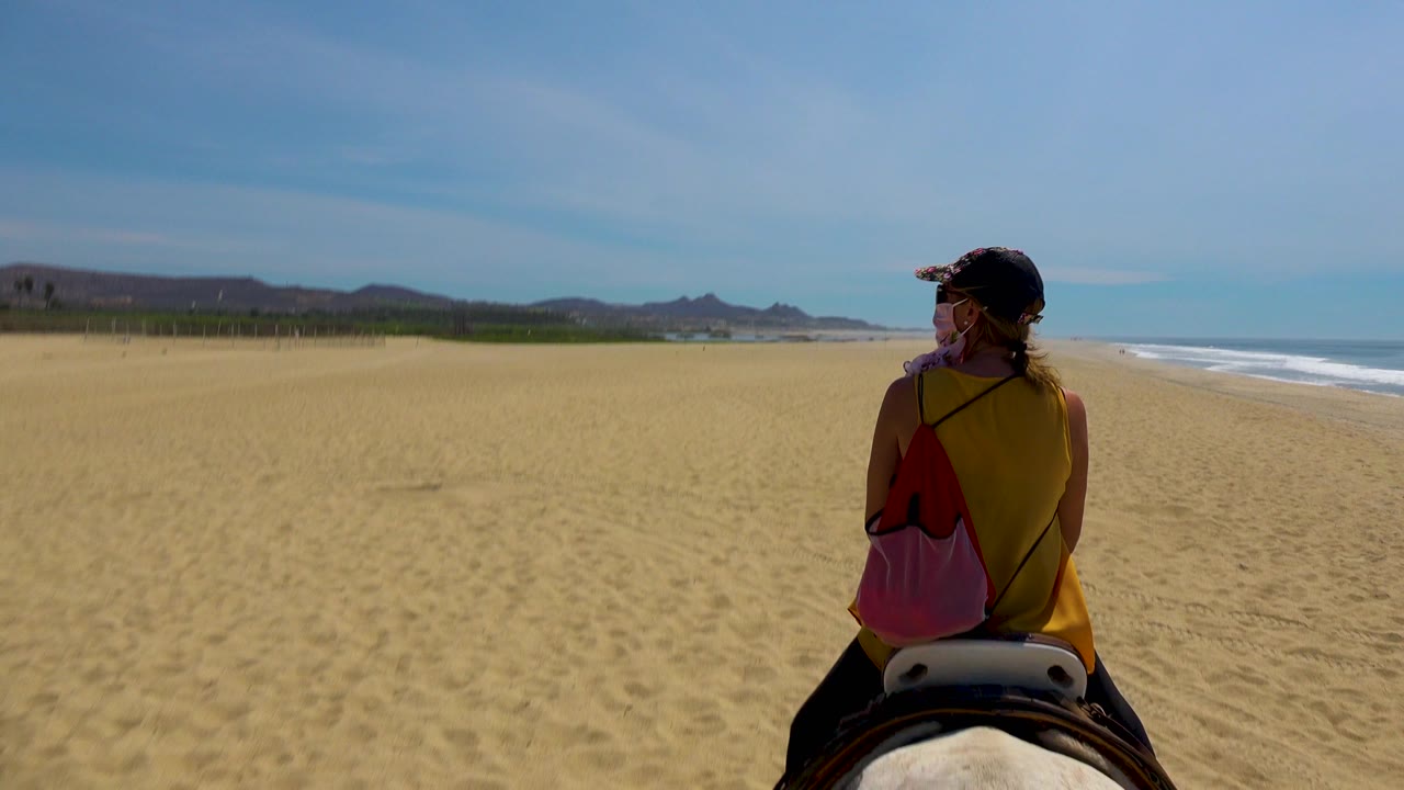 A woman seen from behind rides a horse across a wide sandy beach in San Jose Del Cabo, Mexico, with surf on the right and low hills in the distance.