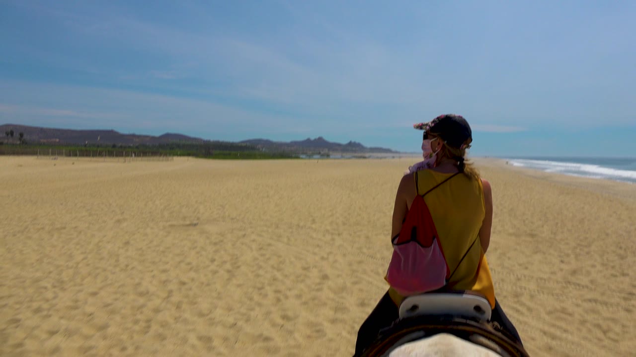 The rider remains centered from behind on the San Jose Del Cabo beach, with a broad stretch of sand ahead and small white waves along the right edge.