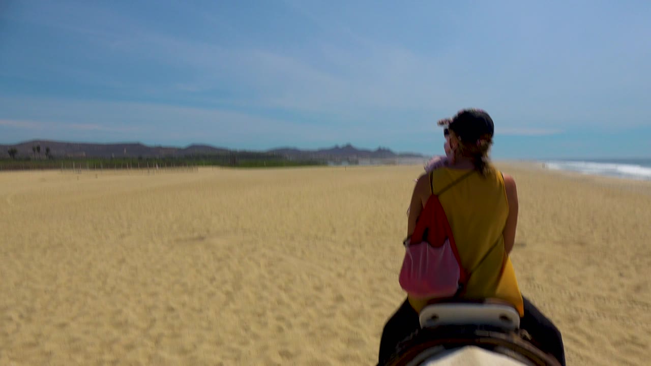 A back view of the horseback rider fills the right side of the frame, with rippled sand, ocean surf, blue sky, and inland hills visible around her.
