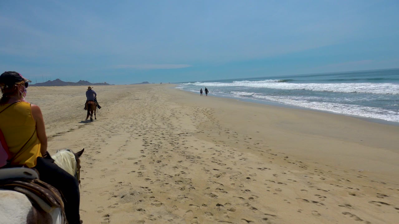 In San Jose Del Cabo, the foreground rider sits partly left of center on a white horse while another rider continues ahead and waves break along the right side.