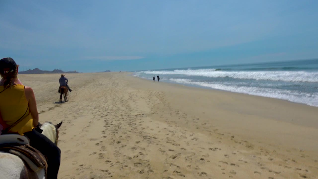 The foreground rider in a yellow top is more centered in the San Jose Del Cabo beach frame, with the leading rider ahead and two small figures walking near the waterline.