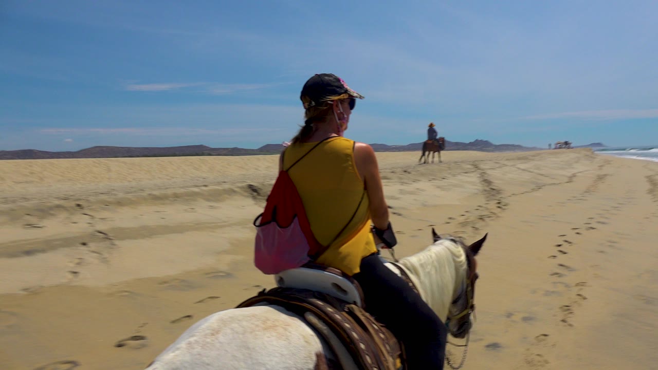 At 0:00 in San Jose del Cabo, Mexico, a foreground rider on a white horse faces down a wide sandy beach toward another rider near the center distance, with waves along the right edge.
