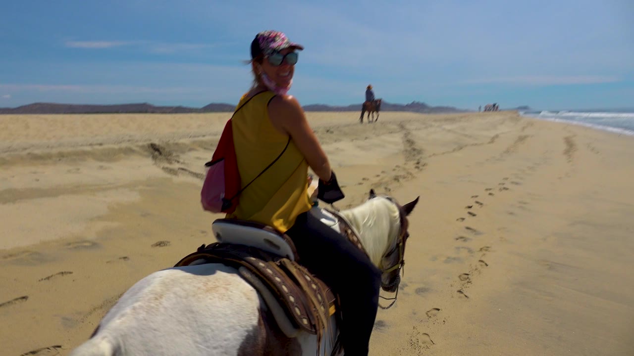 At 0:02 in San Jose del Cabo, Mexico, the foreground rider turns partly toward the camera on the white horse, with a lead rider ahead, footprints and hoof tracks in the sand, and ocean surf to the right.