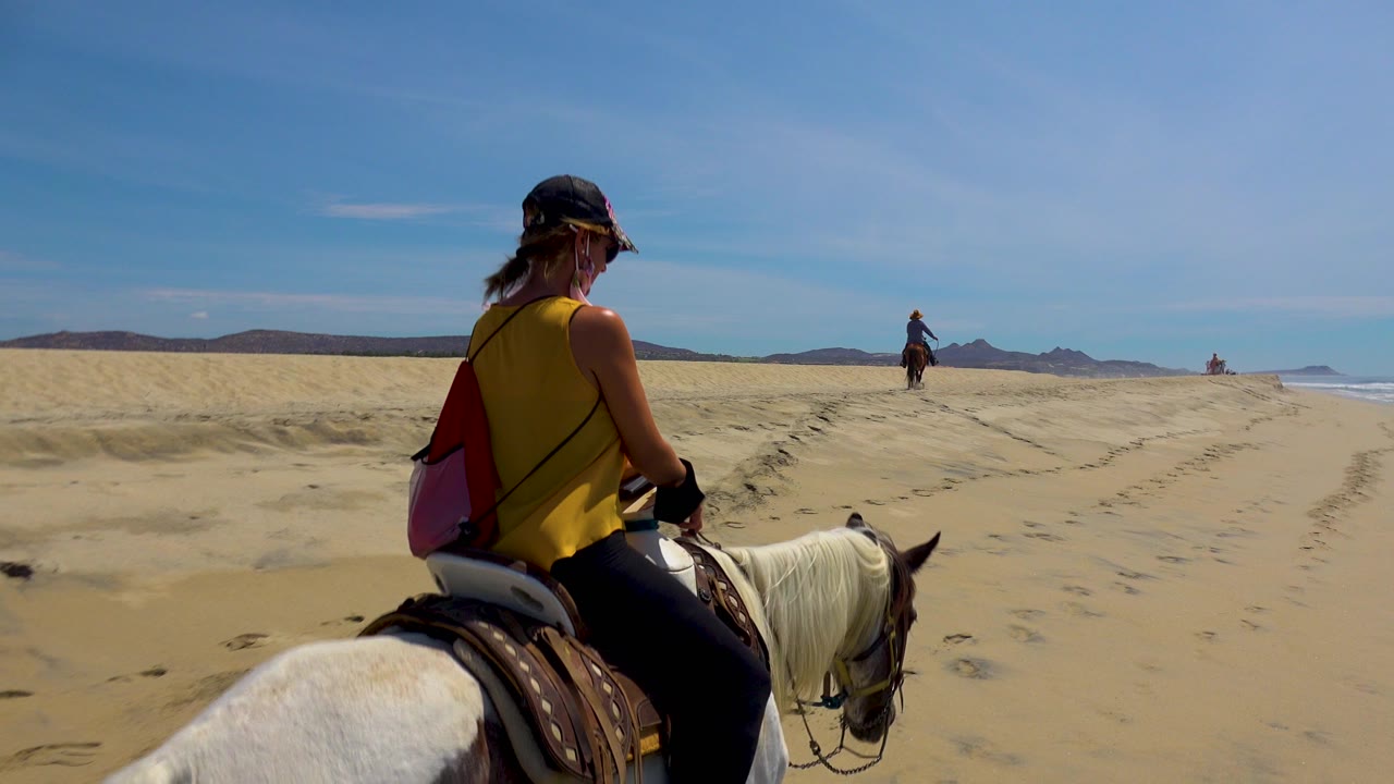 In San Jose Del Cabo, Mexico, a woman in a yellow top rides a white horse across wide sand with another rider in the distance and low hills beyond.