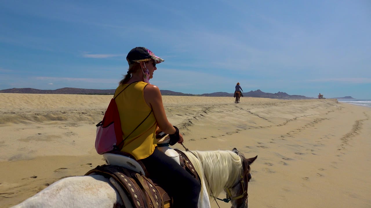 The rider on the white horse remains in the foreground while San Jose Del Cabo beach tracks curve ahead and the ocean line appears on the right side of frame.