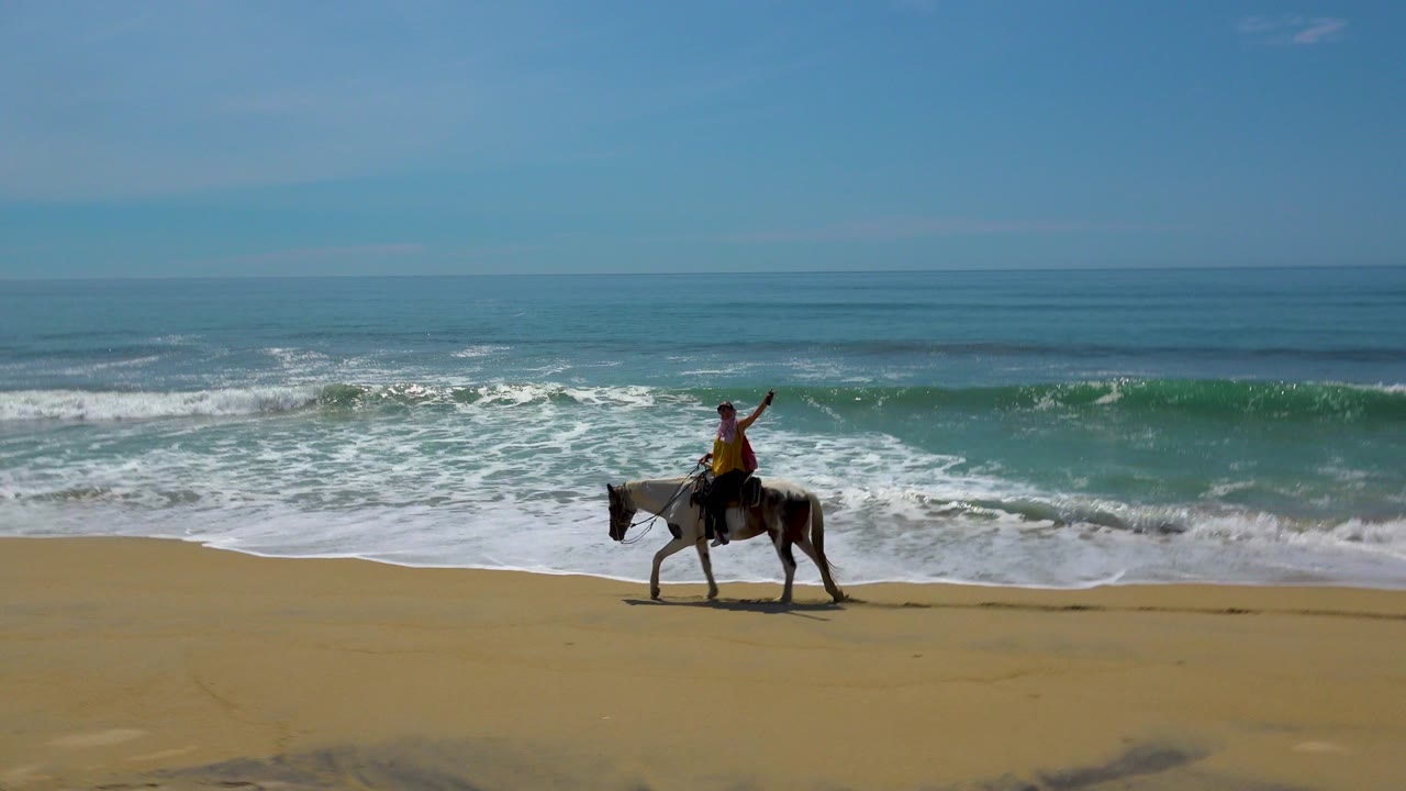 A young woman on horseback rides across sand in San Jose Del Cabo, Mexico, with blue sea and white surf behind her at the start of the clip.