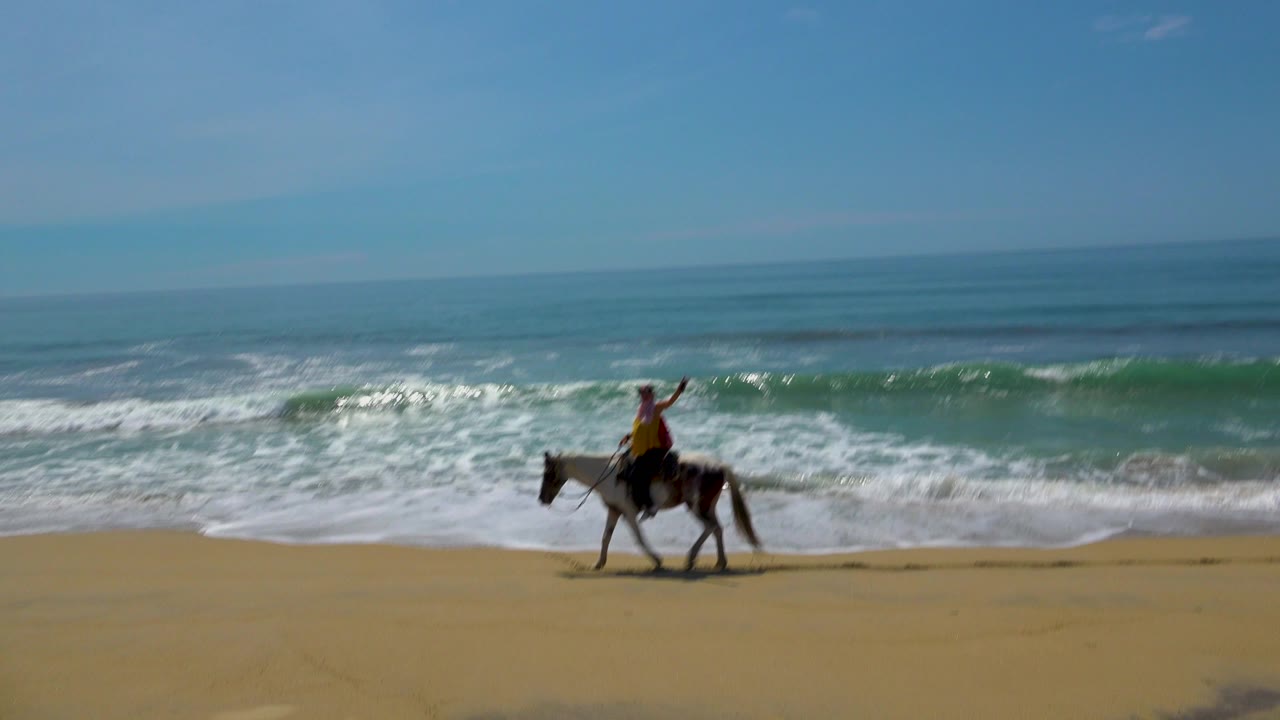 A young woman on horseback appears farther right in the frame, raising one arm while riding beside white surf in San Jose Del Cabo, Mexico.