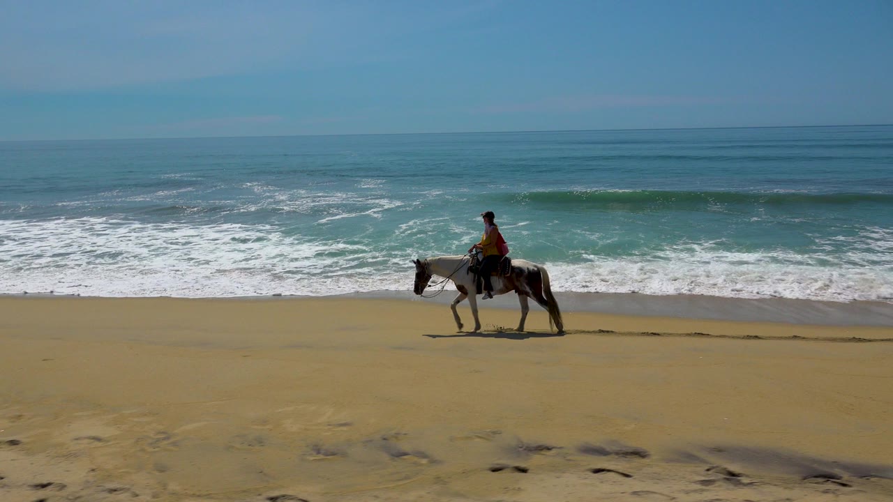 A man rides a horse along the sand in San Jose Del Cabo, Mexico, with white surf rolling behind him and footprints in the foreground.