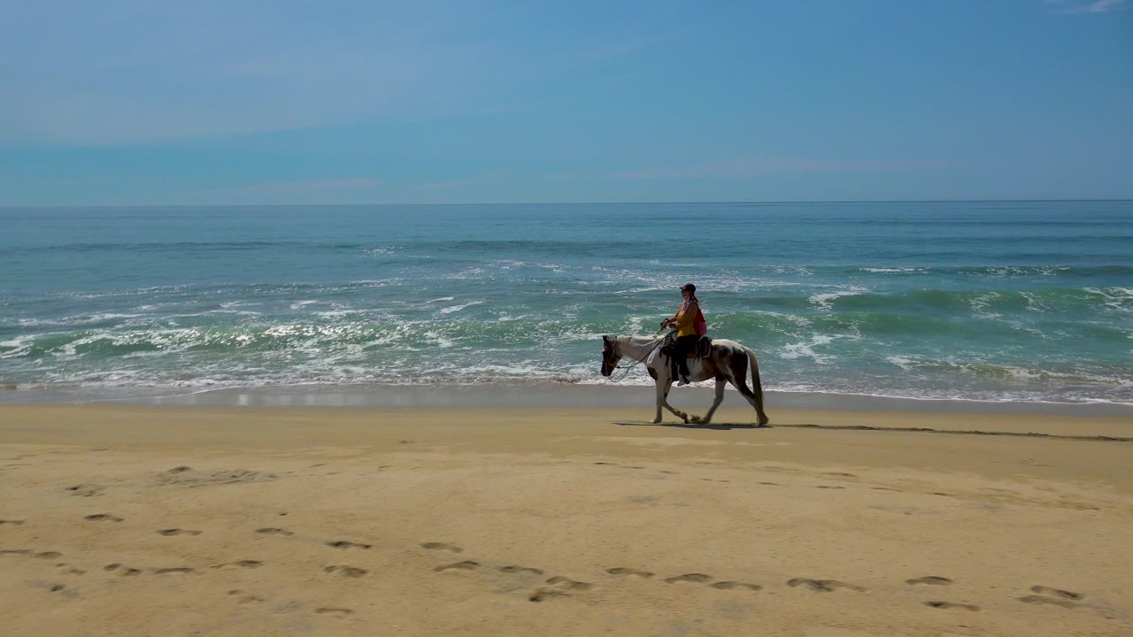 A rider on horseback moves toward the right side of the San Jose Del Cabo beach frame, with a broad strip of empty sand and layered surf behind.