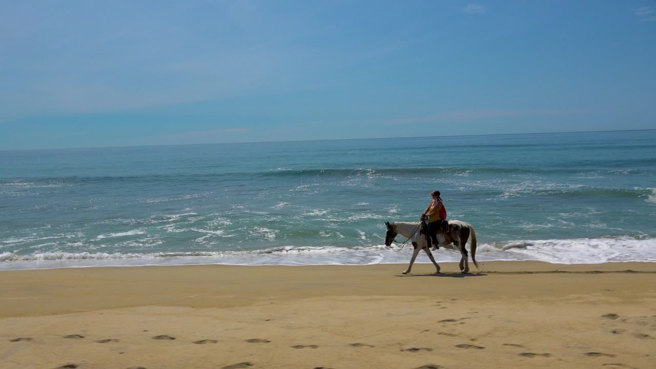 Wide frame from San Jose Del Cabo, Mexico, showing a distant woman riding a spotted appaloosa across tan sand with low surf and blue sky behind her.