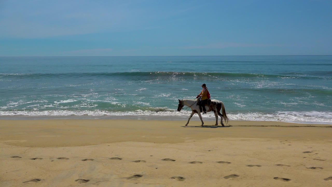 The horse and rider appear farther left in the San Jose Del Cabo beach view, with a darker band of rolling surf forming behind them and footprints in the foreground sand.