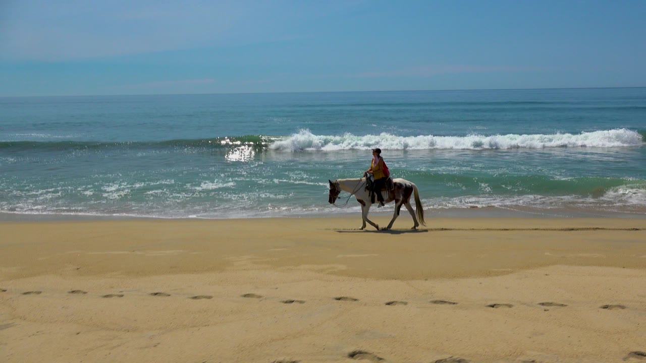 A rider on a horse moves along the sandy beach in San Jose Del Cabo, Mexico, with low surf breaking behind them and a wide blue sky above.