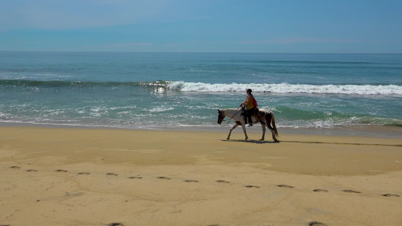 A visible rider on horseback appears near the right side of the San Jose Del Cabo beach frame, with broad empty sand and blue-green water behind.